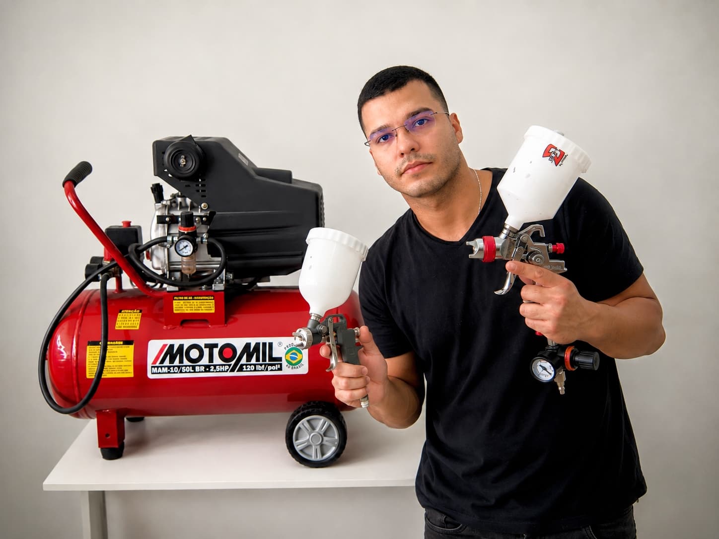 Gabriel Santos segurando duas pistolas de pintura ao lado de um compressor de ar em sua oficina